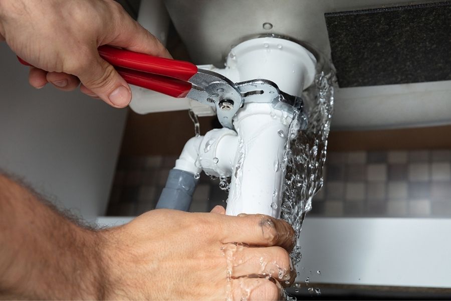 A close-up of a plumber using red-handled pliers to tighten a white PVC pipe under a leaking sink.