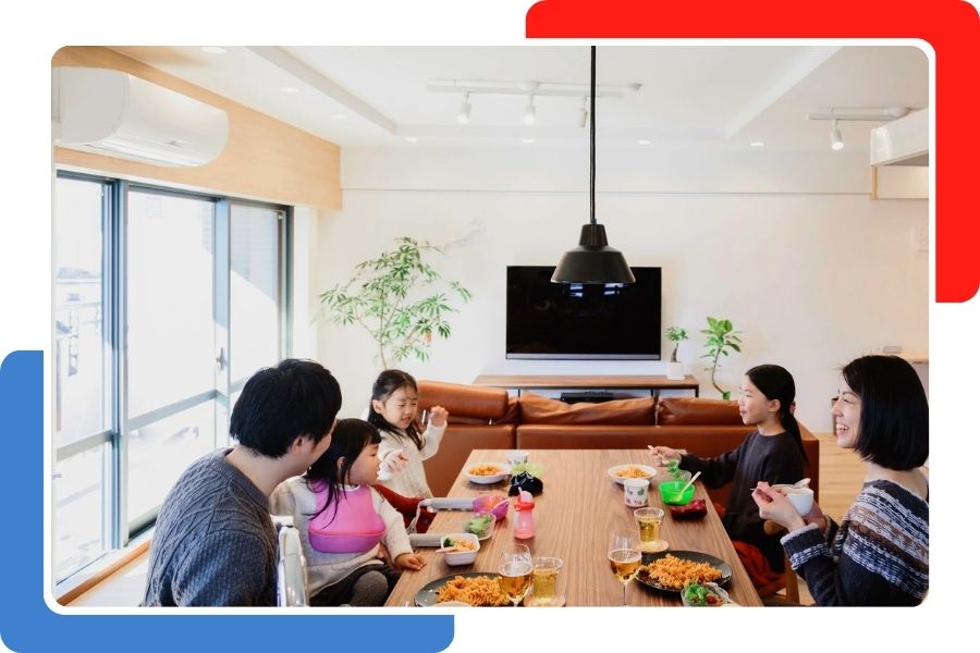 A family of five sitting at a wooden dining table eating a meal, with a white ductless mini-split air conditioner mounted on the wall above the window.