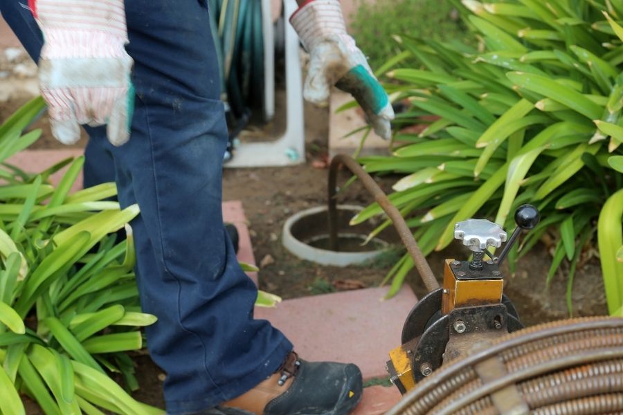 A technician in work gloves and blue uniform operating a motorized drain cleaning machine near an outdoor sewer cleanout.