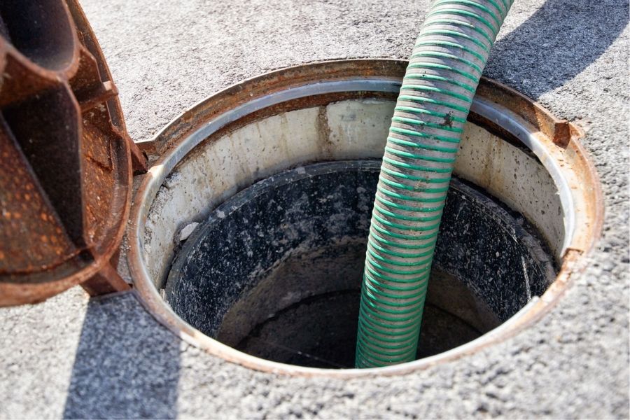 A thick green suction hose inserted into an open concrete manhole on an asphalt surface.