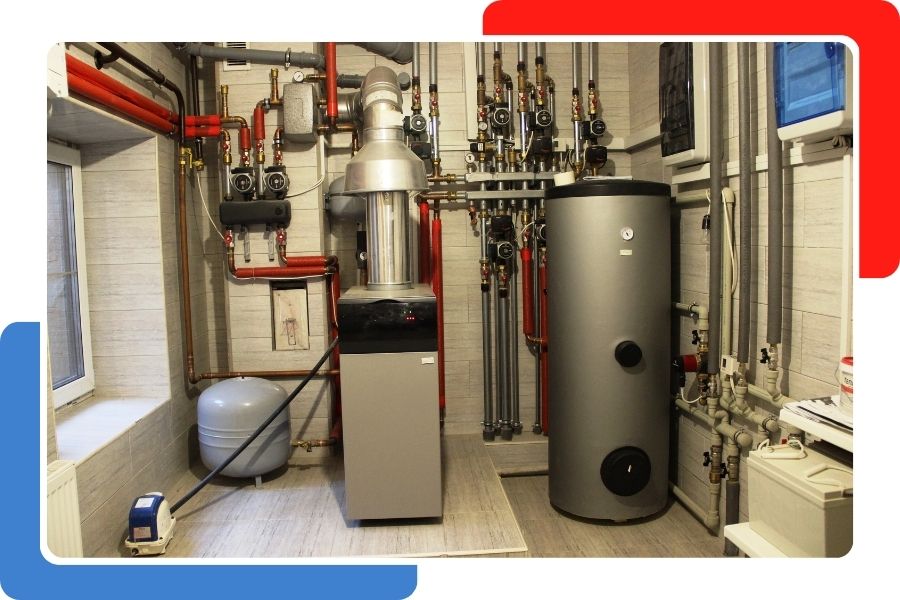 A utility room containing a modern gray furnace, a large vertical water tank, and complex wall-mounted piping with red insulation.